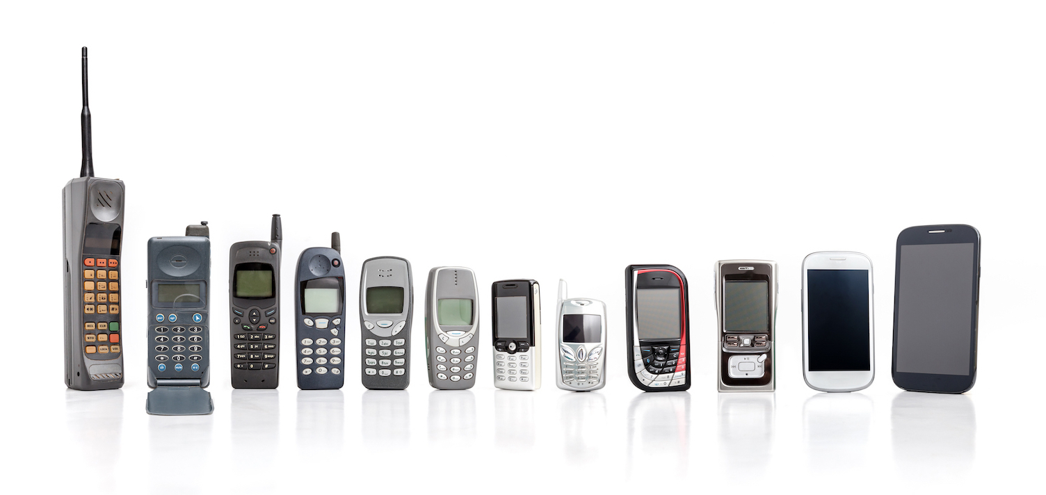 Pho­to­graph show­ing the evol­u­tion of cell phones from 1973 to 2015. From left to right there is a large hand­held phone with a long aer­i­al, a large dial pad and a small screen, fol­lowed by mod­els de­creas­ing in size, through to the smart phones which are slightly big­ger with full-size screens and no dial pads.